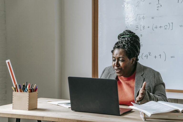 African American female teacher talking during class on netbook while sitting at desk with books and stationery in classroom